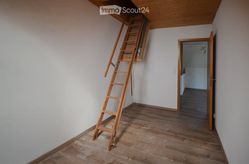 Empty room with wooden flooring, white walls, and a wooden ladder leading to an attic space. An open doorway leads to another room with similar flooring.