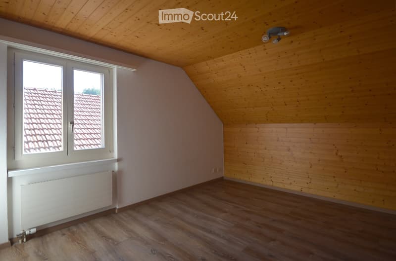 Empty attic room with wooden walls, wooden floor, large window showing a roof view, and a radiator under the window.
