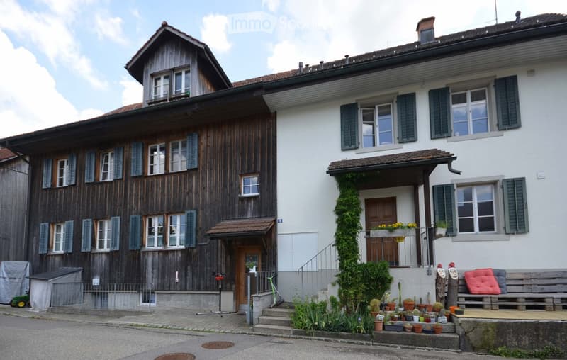 Two-story house with a wooden facade on the left and a white facade on the right, featuring multiple windows with shutters, a small porch, and a garden area with potted plants and a bench.