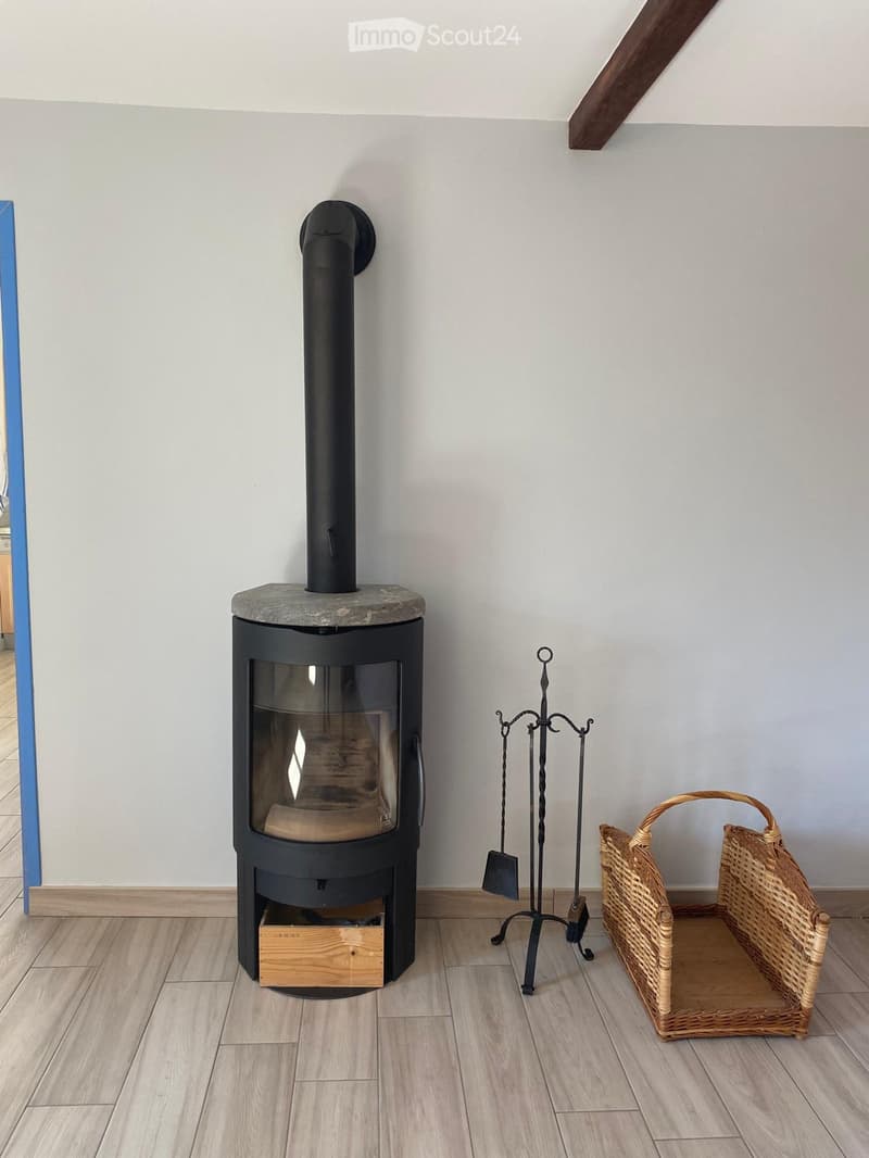 Modern wood-burning stove with a black chimney, surrounded by fireplace tools and a wicker basket on a light wooden floor.