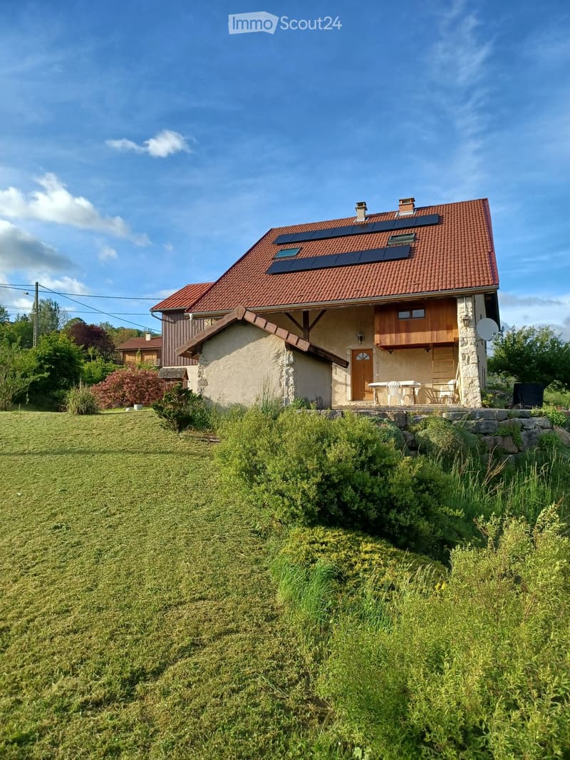Two-story house with red-tiled roof, solar panels, stone and wood exterior, small balcony with outdoor furniture, surrounded by greenery.