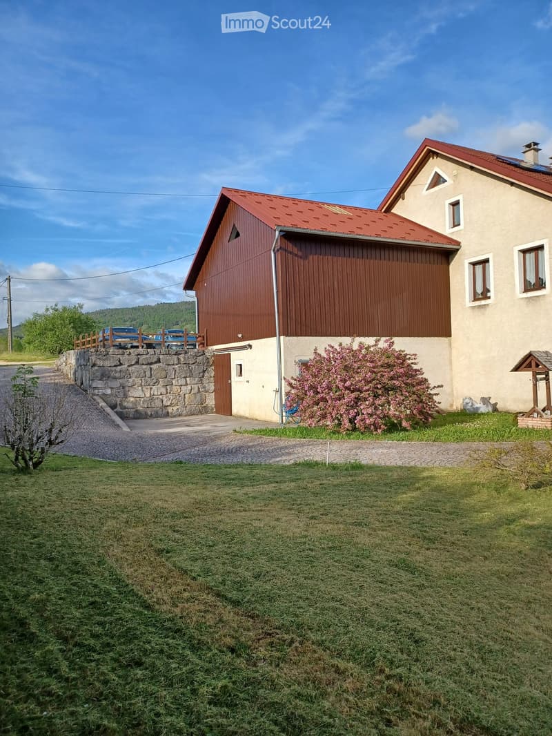 Two-story house with red-tiled roof, beige walls, and a wooden garage. Features a stone retaining wall, driveway, and landscaped garden with flowering shrubs and a small wooden structure.
