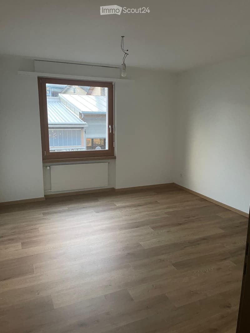 Empty room with wooden flooring, white walls, and a large window with a view of a rooftop area.