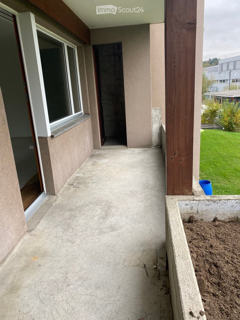Concrete balcony with wooden railing, large window, and view of a grassy yard.