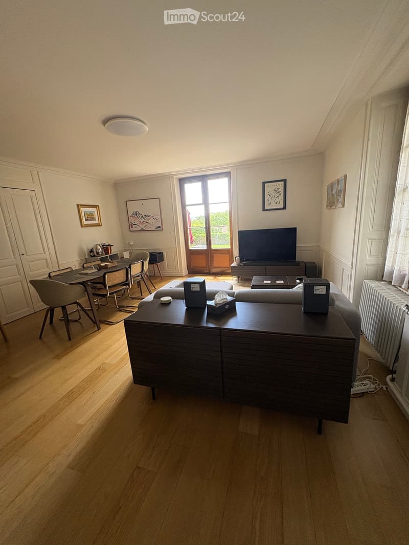 A modern living room with wooden flooring, a grey sofa, a coffee table, a dining area with chairs and a table, a television, and several picture frames on the walls.