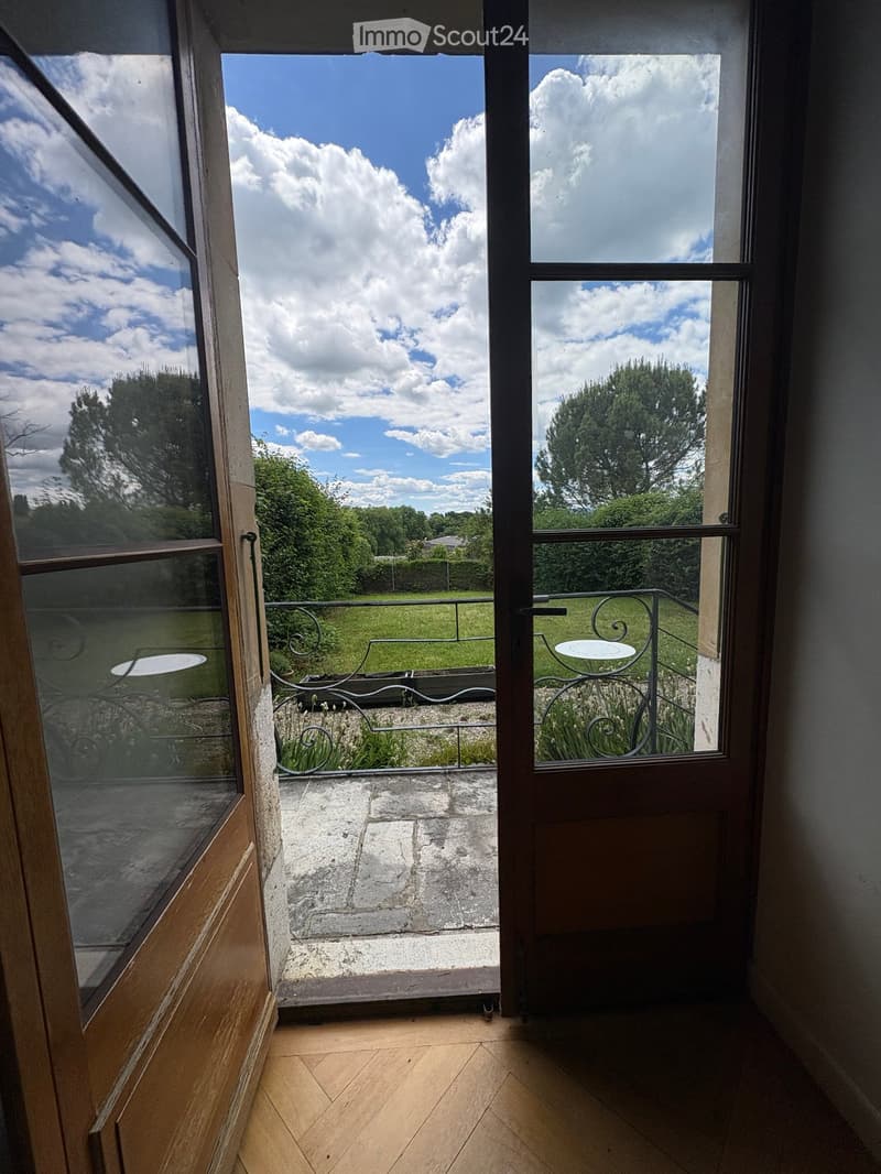 View from inside a room through a large open doorway onto a stone-paved balcony with metal railings, small tables, and a view of a green lawn and trees under a partly cloudy sky.