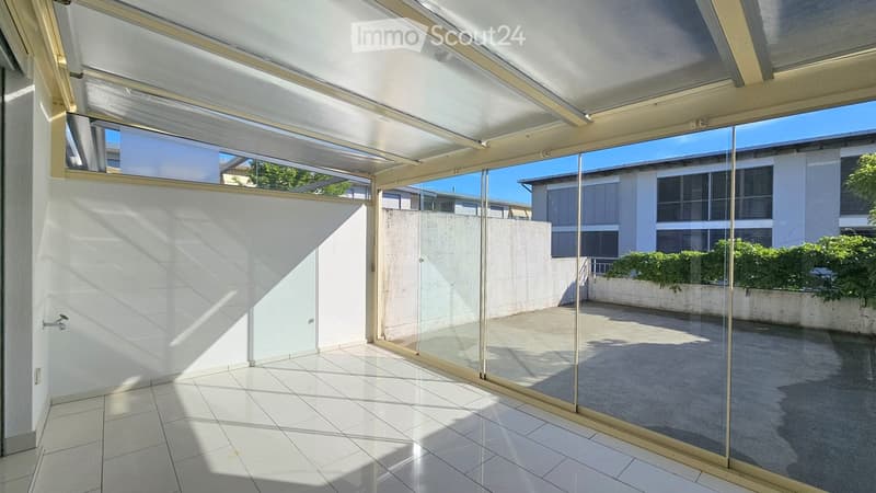 Bright, enclosed balcony with white tiled flooring, glass walls, and a retractable roof, offering views of a modern building and greenery.