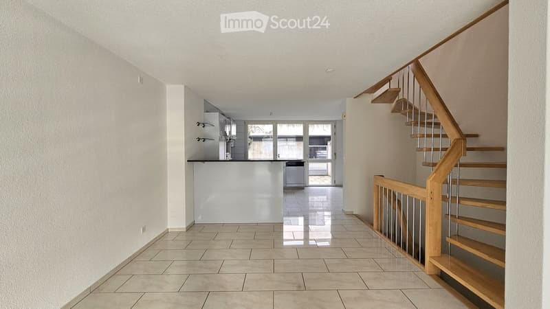 Bright hallway with tiled floor, wooden staircase with metal railing, and an open kitchen area visible in the background.