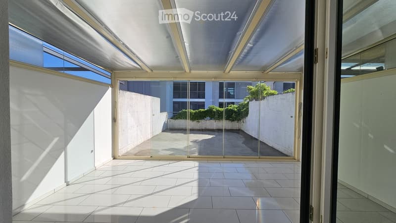 Bright, open terrace with white tiled flooring, large glass sliding doors, and a view of a neighboring building and greenery.