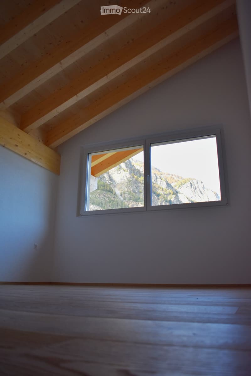 A room with wooden beams, a window with a mountain view, a white wall, and a wooden floor