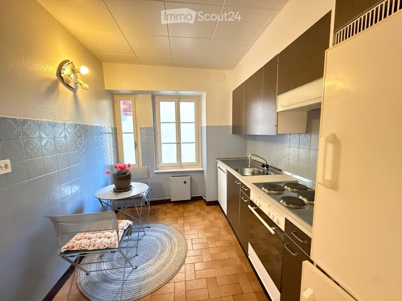 A compact kitchen with modern cabinets, a small dining area, and a round rug. Features include a sink, stove, refrigerator, and a small table with a chair.