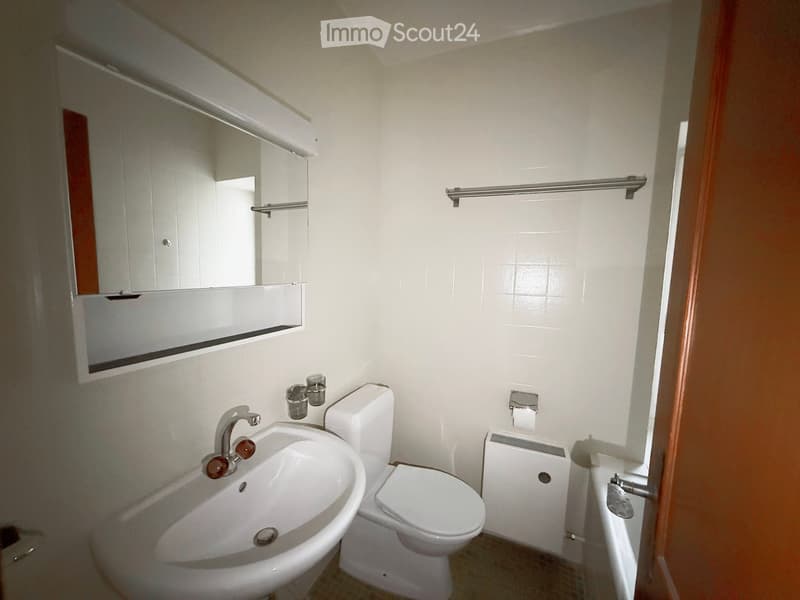 A small bathroom with a white sink, toilet, mirror, and towel rack. The walls are tiled, and there is a small window on the right side.