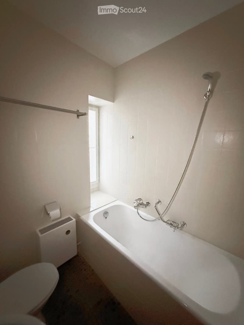 Bathroom with a white bathtub, faucet, toilet, radiator, and towel rack. The room has tiled walls and a small window letting in natural light.