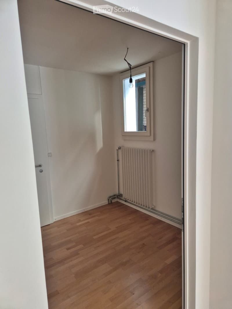 Empty room with wooden flooring, white walls, a radiator, and a small window with a mirror above it.