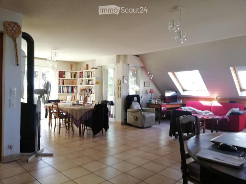 A spacious living room with a red sofa, a beige armchair, a dining table with chairs, a bookshelf, and a laptop on a small table. The room features large windows and skylights, with a tiled floor and modern lighting fixtures.