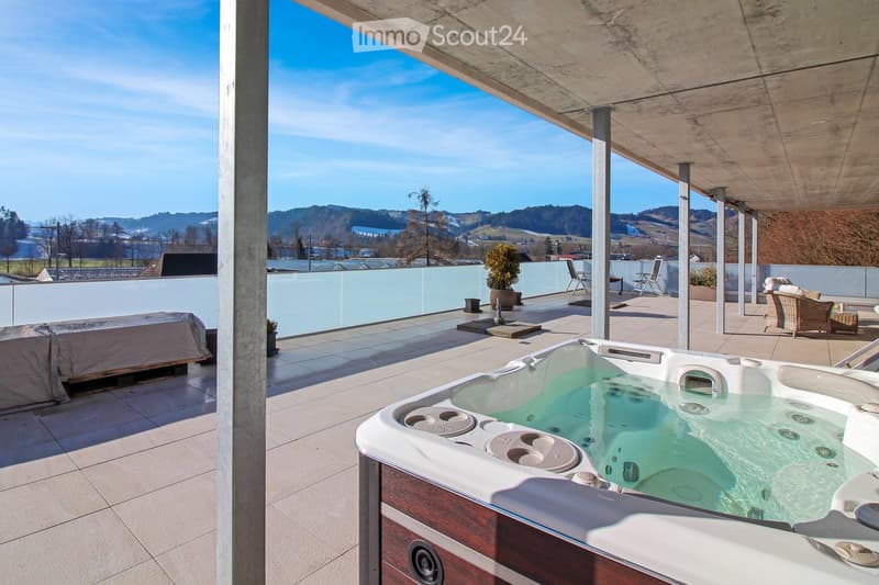 Modern rooftop terrace with a hot tub, lounge chairs, a small table, and potted plants, offering scenic views of the surrounding landscape.