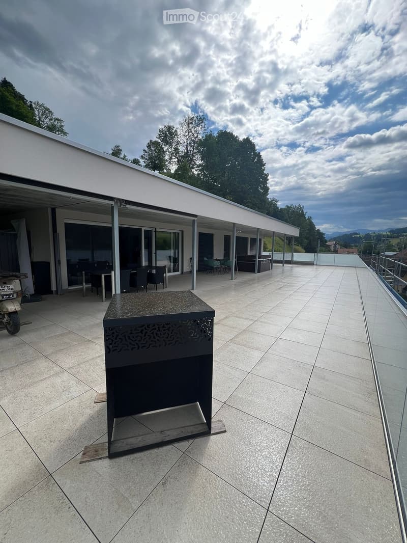 Modern outdoor patio with tiled flooring, glass railing, and a covered seating area with tables and chairs. A black barbecue grill is placed on the patio.