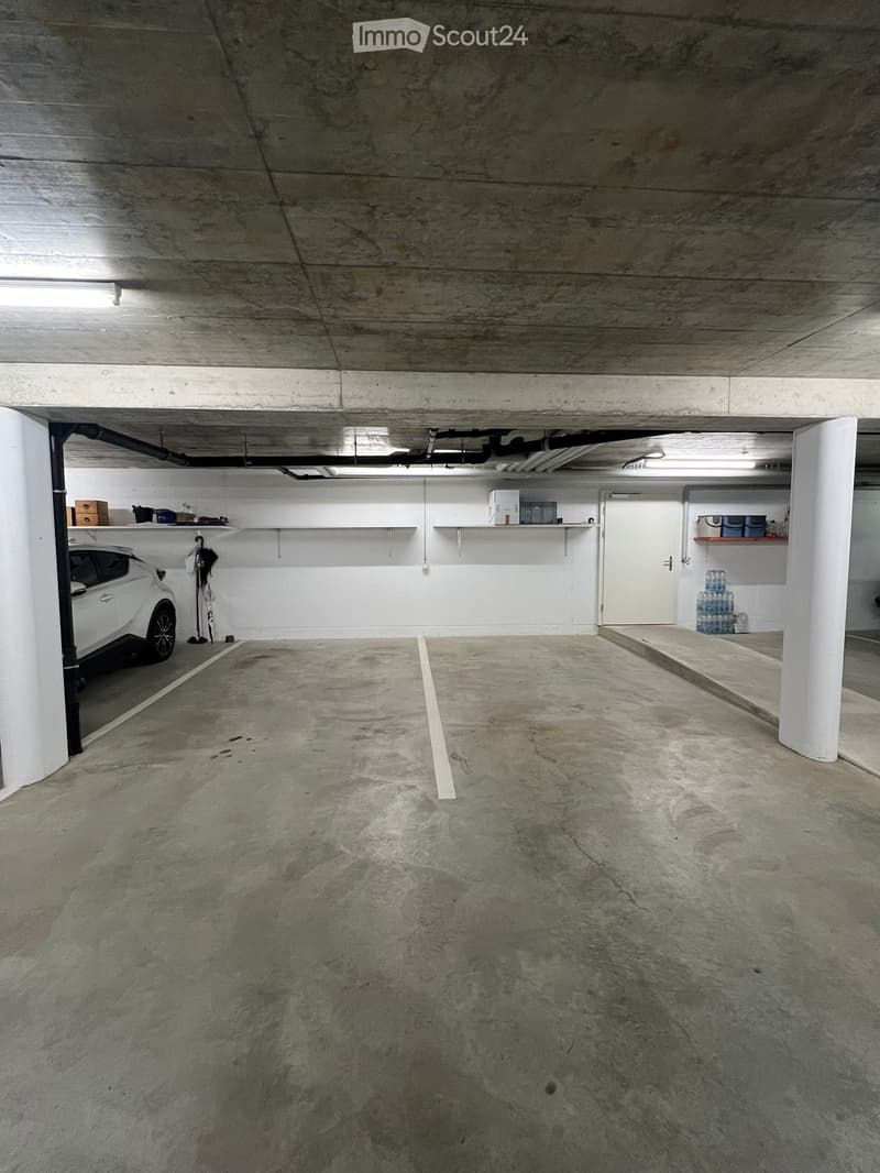 Underground parking space with a white car parked on the left, shelves with boxes and items, and a concrete floor.