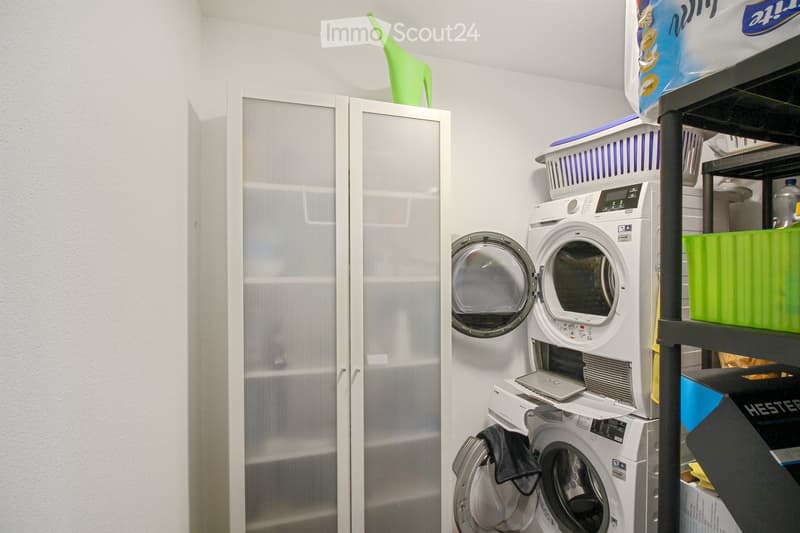 A compact bathroom with a washing machine and dryer stacked vertically, a mirrored cabinet, and a shelf with laundry baskets and cleaning supplies.