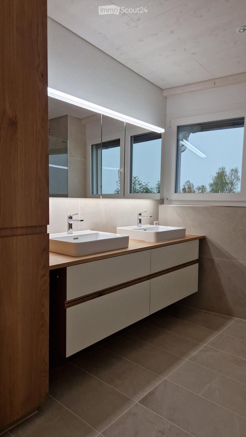 Modern bathroom with two white sinks on a wooden countertop, sleek faucets, and built-in cabinets. Large windows provide natural light.