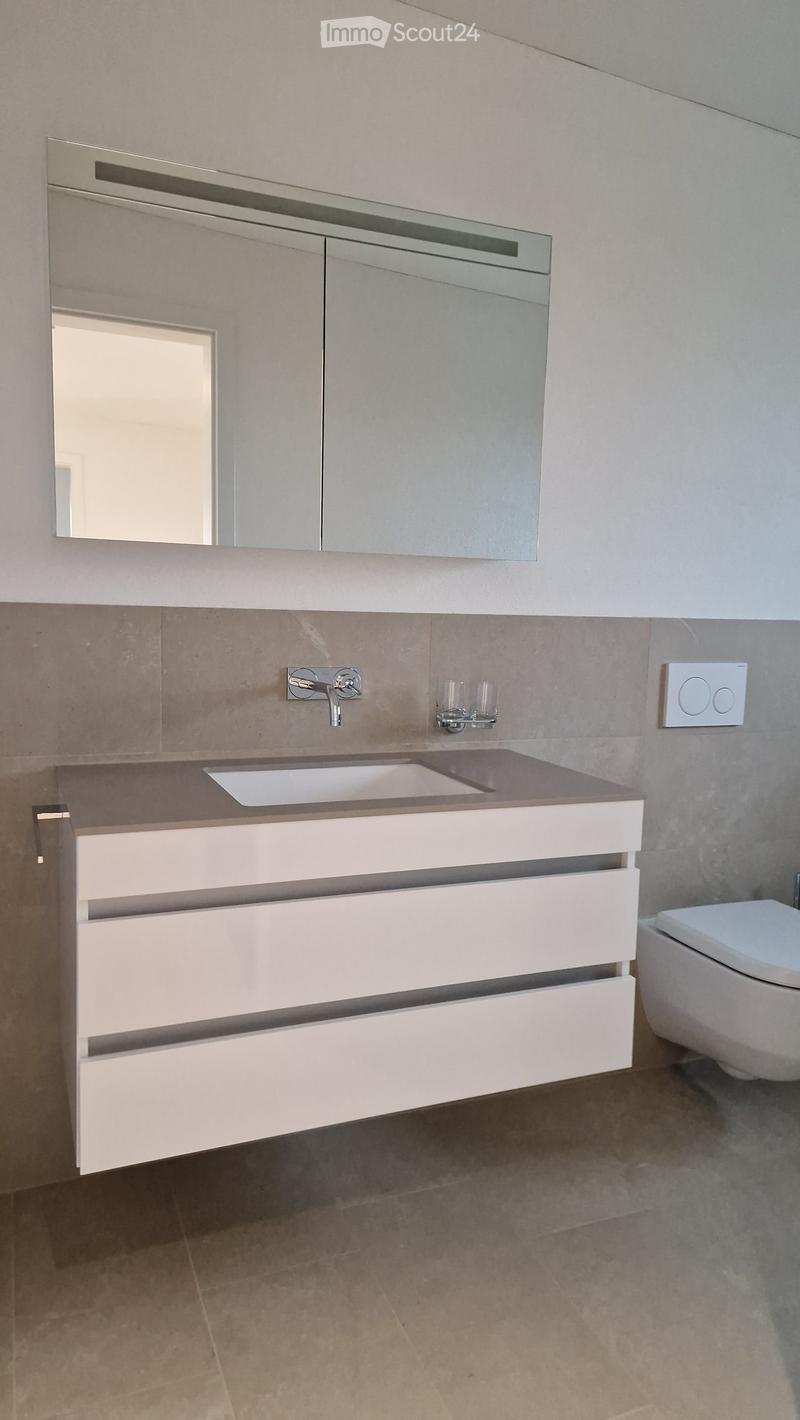 Modern bathroom with a white vanity, sink, mirror, and toilet. The vanity has two drawers and a countertop with a built-in sink. A modern faucet is mounted above the sink, and a mirror with a sleek frame is mounted above the vanity. Two small cups are pla