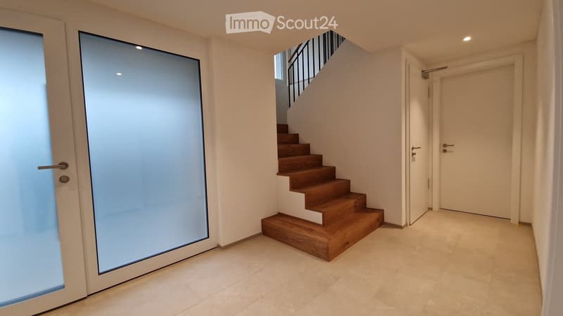 Modern hallway with a wooden staircase leading upwards, white walls, tiled floor, and a glass door on the left.