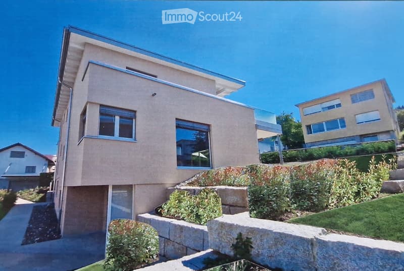 Modern two-story house with beige facade, large windows, and a raised ground floor. The house features a sleek design with a glass balcony on the upper level and is surrounded by landscaped greenery and stone steps.