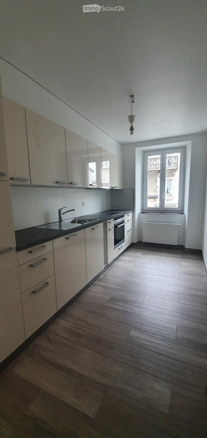 Empty kitchen with white cabinets, dark countertops, and wooden flooring. Features a sink, faucet, and a window with decorative ironwork.