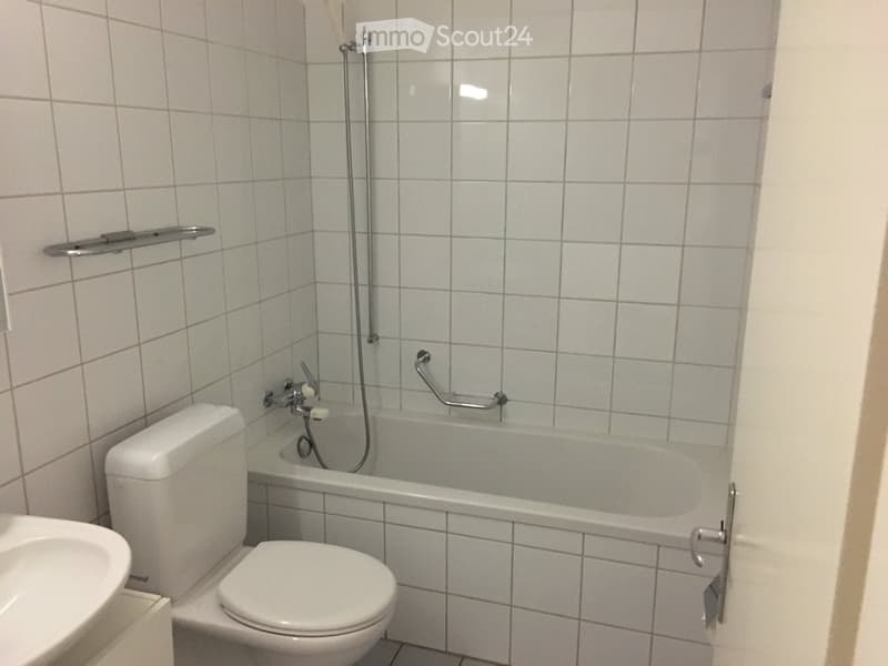 A small bathroom with white tiled walls and floor, featuring a bathtub with a shower handle, a toilet, and a towel rack on the wall.