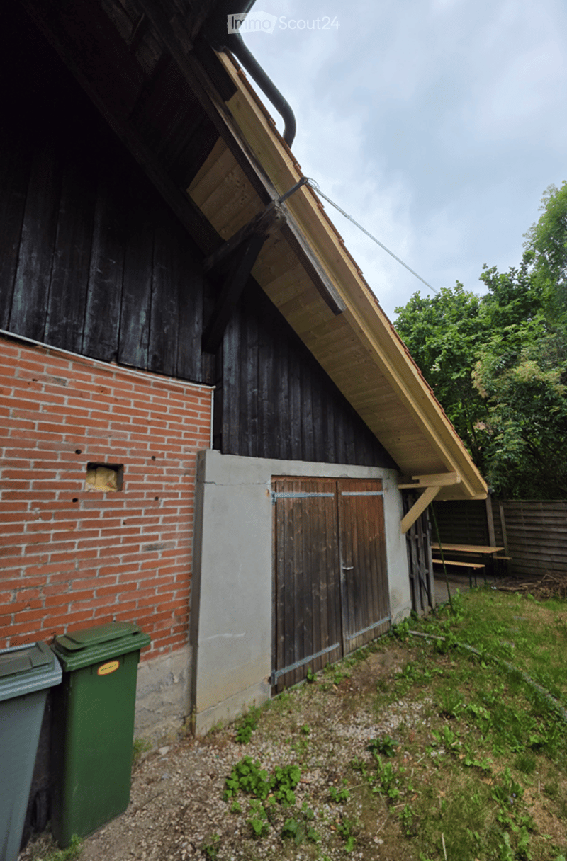 A rustic wooden barn with a brick wall section, featuring a large wooden door and a slanted roof. Green trash bins are placed in front of the barn, and a wooden fence and trees are visible in the background.