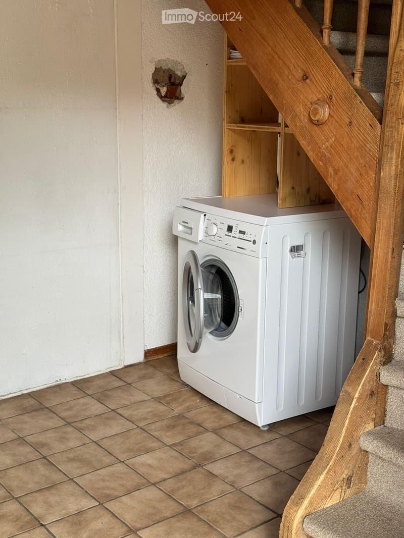 A small bathroom with a white washing machine placed next to a wooden staircase. The floor is tiled, and the walls are painted in a light color with a visible hole.