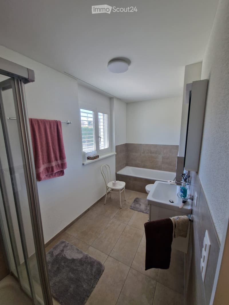 Modern bathroom with a bathtub, sink, toilet, and a small chair. Features tiled walls, a window, and towels on the floor.