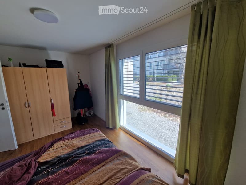 Bedroom with wooden floor, large window with blinds, bed with patterned bedspread, wardrobe, and curtain.