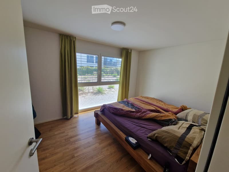 A small bedroom with a wooden floor, a bed with a purple mattress and patterned blankets, and a window with green curtains.