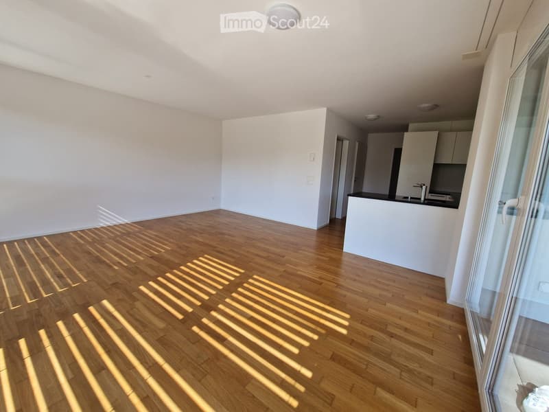 Empty room with wooden flooring and white walls, featuring a small kitchen area visible in the background.