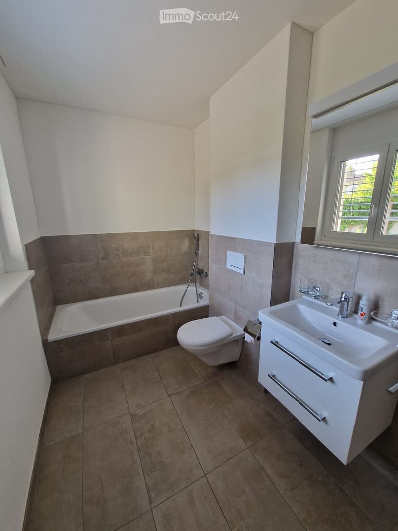 Modern bathroom with a bathtub, toilet, sink, and mirror. Brown tiles on walls and floor, white fixtures, and a window providing natural light.