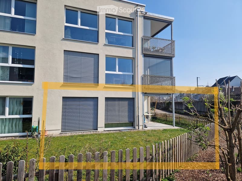 Modern two-story house with large windows, balcony, and a well-maintained garden with a wooden fence.