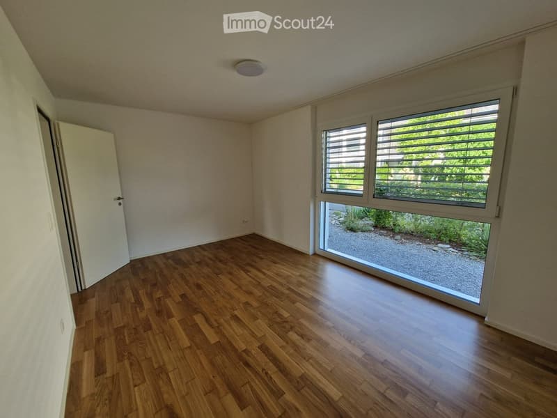 Empty room with wooden flooring, white walls, and large windows with blinds overlooking a garden area.