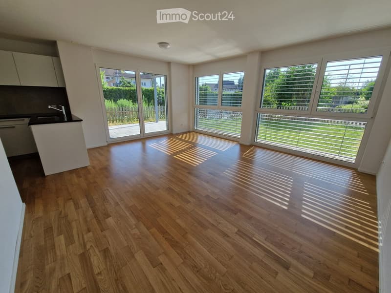 Empty room with wooden flooring, large windows, and a small kitchen area visible on the left side.