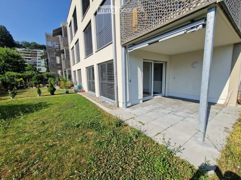 A modern house with a well-maintained garden, featuring a paved pathway leading to a covered entrance area with large windows and sliding doors. The garden includes neatly trimmed grass, small shrubs, and a few potted plants.