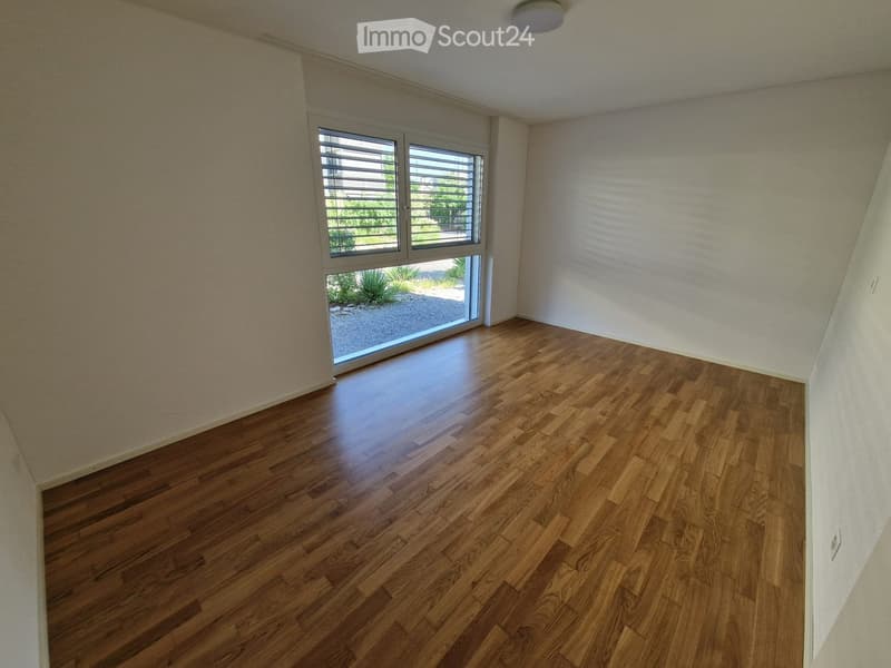 Empty room with wooden flooring, white walls, and large windows with blinds, offering a view of the outdoor area.