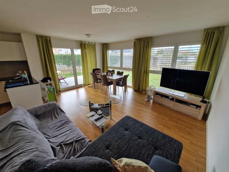 Open-plan living and dining area with wooden flooring, large windows, a sofa, coffee table, TV, dining table with chairs, and a fan.