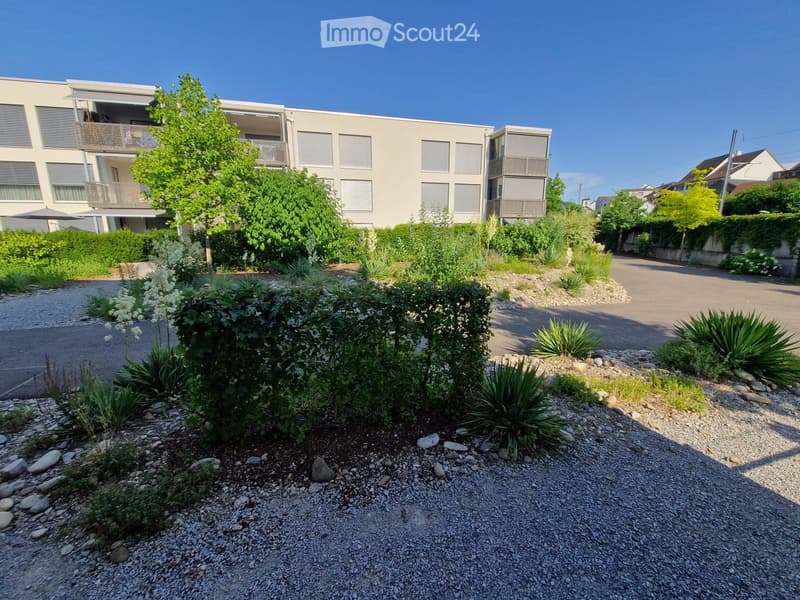 Modern residential building with balconies, surrounded by landscaped garden area with gravel pathways, shrubs, and small trees.