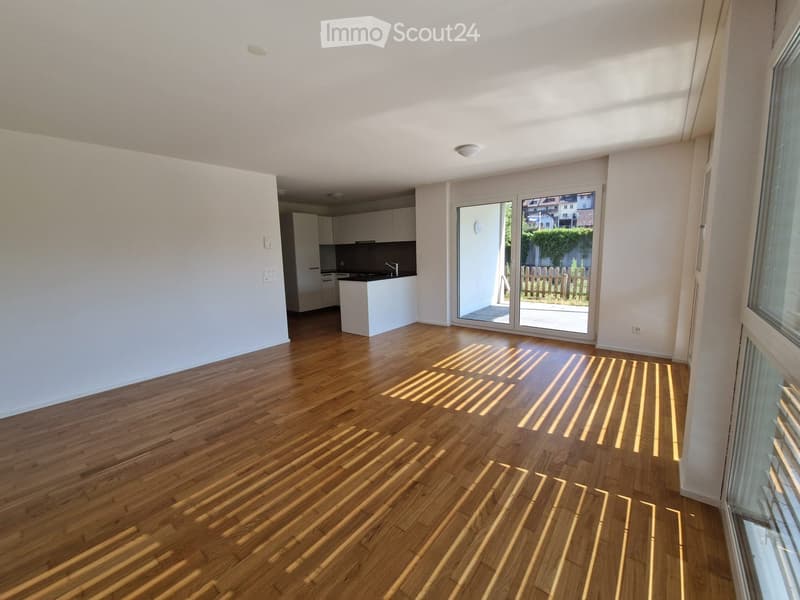 Bright, unfurnished living space with wooden flooring, large sliding glass doors leading to a patio, and an adjacent kitchen area with white cabinets and a countertop.