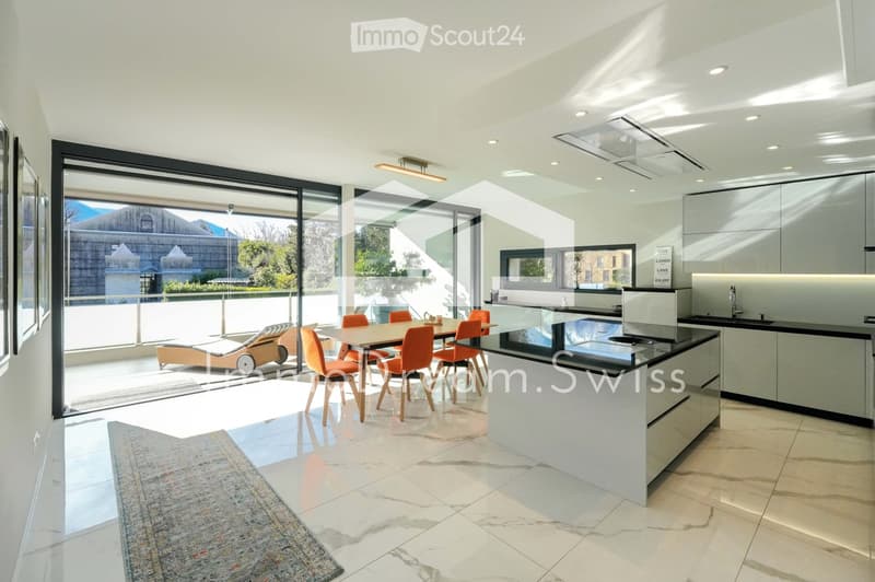 Modern, open-plan kitchen and dining area with sleek white cabinets, a central island, and a dining table with orange chairs. Large glass windows offer a scenic view and access to a sun lounger on the balcony.