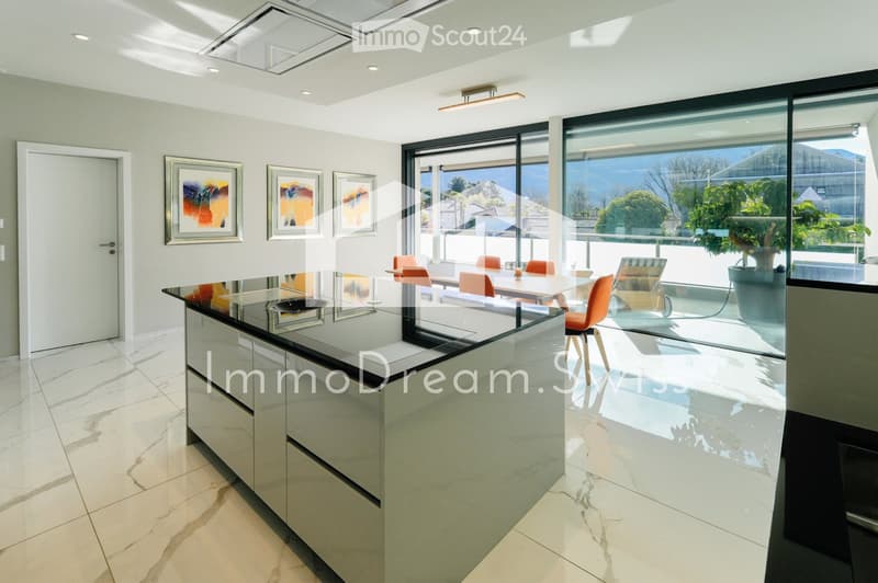Modern kitchen with a glass island, white cabinets, and a dining area with orange chairs and a table, overlooking a scenic view through large windows.