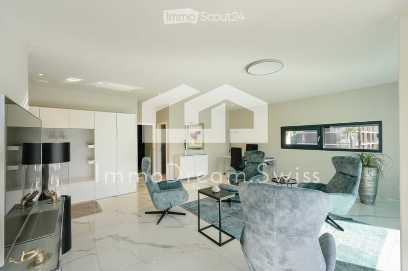 Modern living room with light-colored walls, white cabinets, and large windows. Features include a grey sofa, armchairs, a coffee table, a floor lamp, and decorative items.