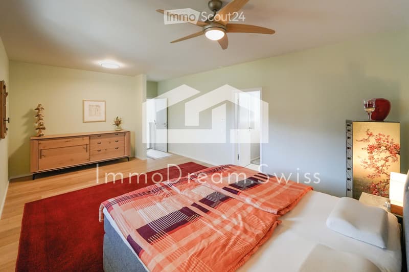 Bright bedroom with wooden flooring, a large bed with a colorful quilt, a wooden dresser, a nightstand with a lamp, a ceiling fan, and a small decorative table with a vase.