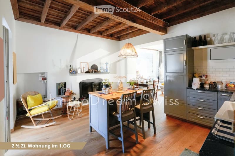 A cozy, well-furnished room with a kitchen area and a living space. Features include a wooden kitchen island with chairs, a rocking chair, a small stool, a fireplace with decorative items, and modern kitchen cabinets.