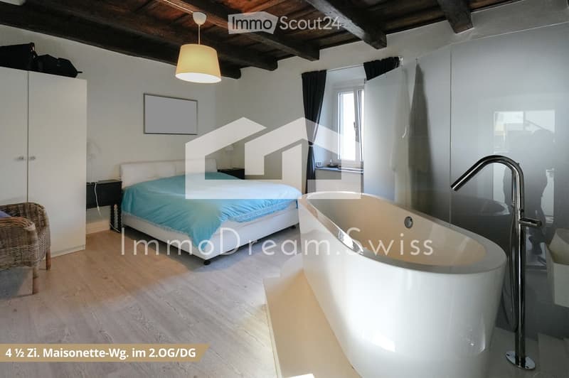 Cozy bedroom with a white bed covered in blue bedding, a wicker chair, a modern bathtub, a large mirror, and a wardrobe. The room features wooden flooring and exposed wooden beams on the ceiling.